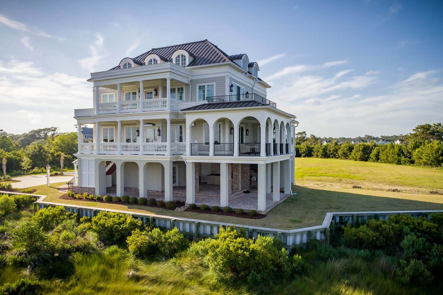 A large, elegant coastal mansion with multiple balconies and a spacious lawn surrounded by greenery.